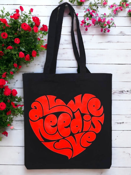 A black tote bag with All we need is love written on it is placed on a simple background. 